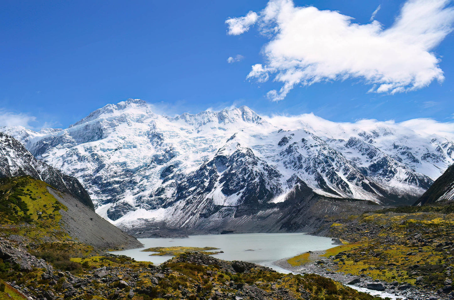 Snow-capped mountains with a lake at their base under a blue sky with clouds.