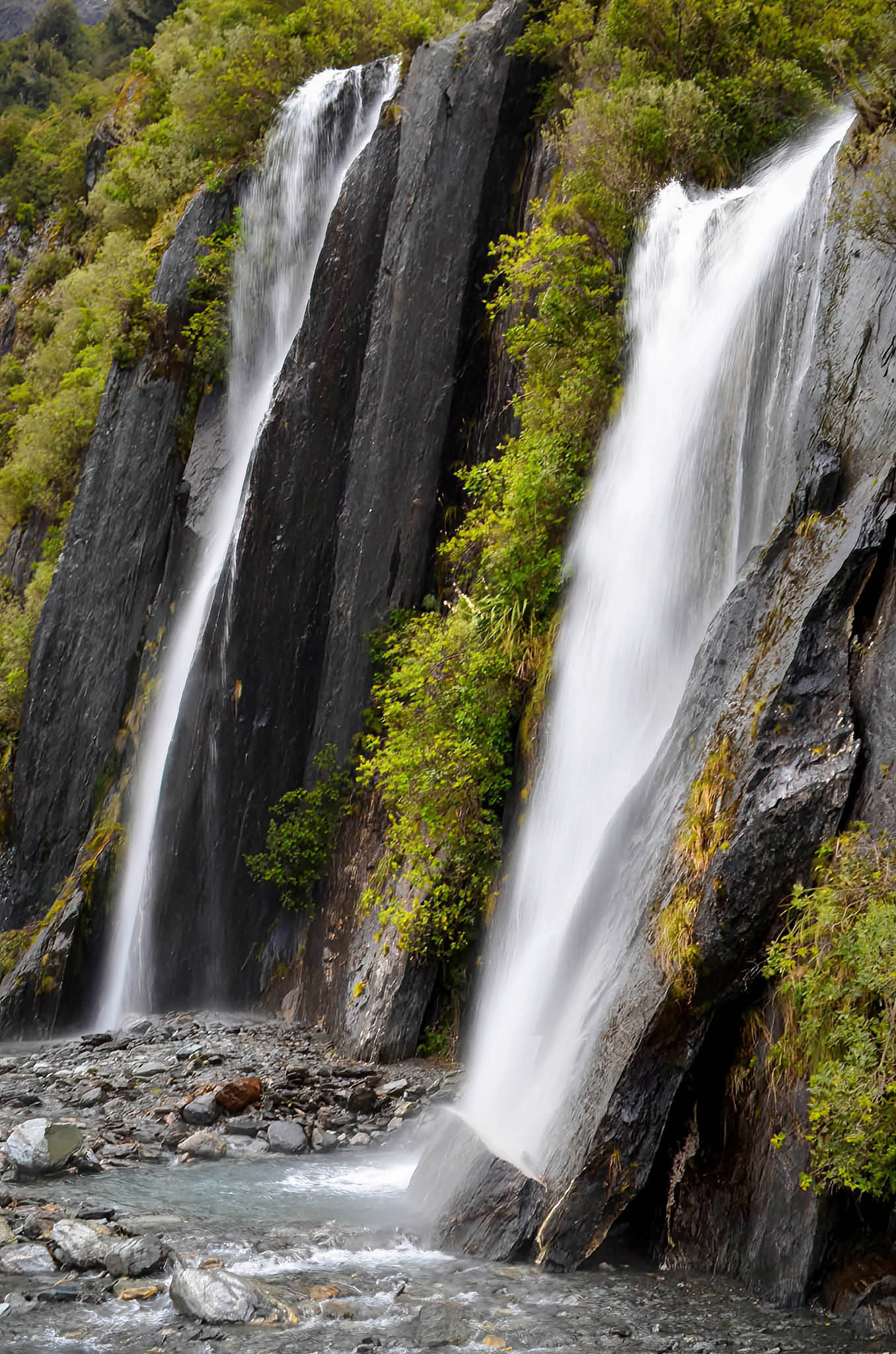 Two waterfalls cascading down rocky cliffs with greenery in a natural setting.
