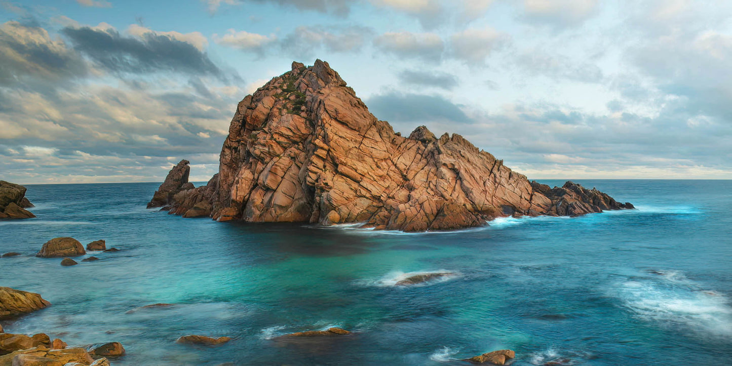 Rugged coastal landscape with large rock formations and blue ocean under a cloudy sky.