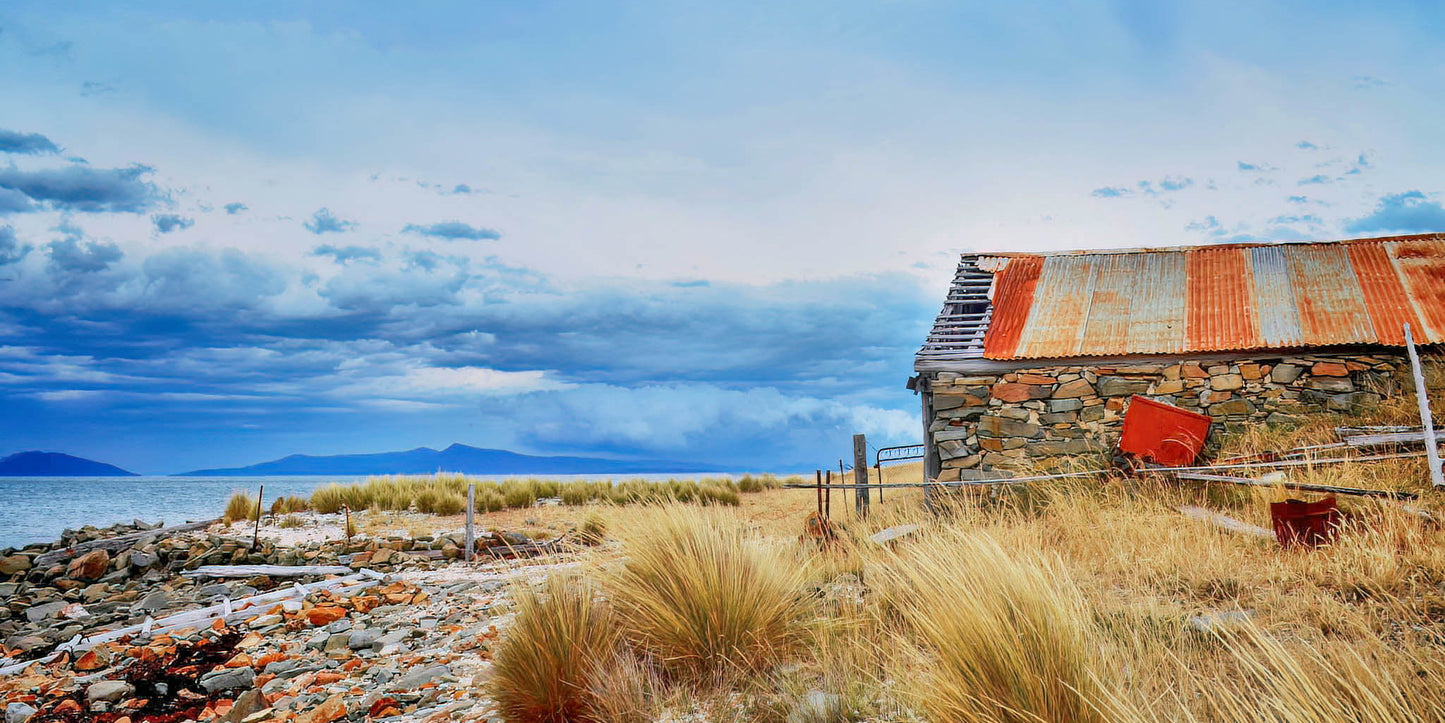 Rustic stone building with a corrugated metal roof near a body of water under a stormy sky with clouds.