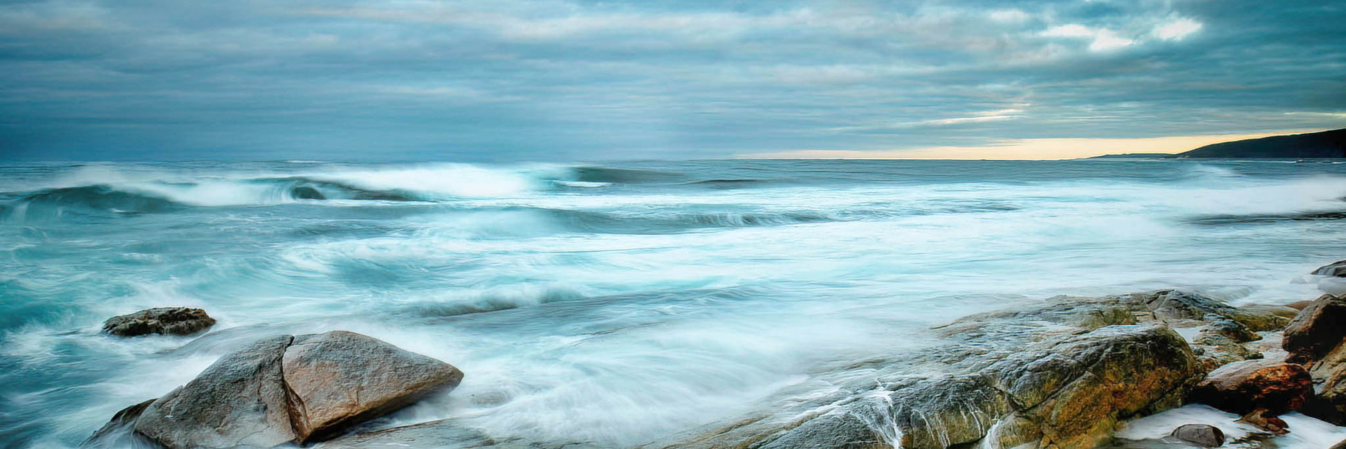 Ocean waves crashing against rocks with a cloudy sky.