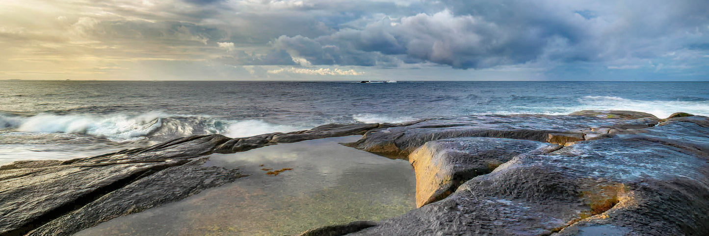 Rock pool on a ledge with ocean waves and a stormy sky.