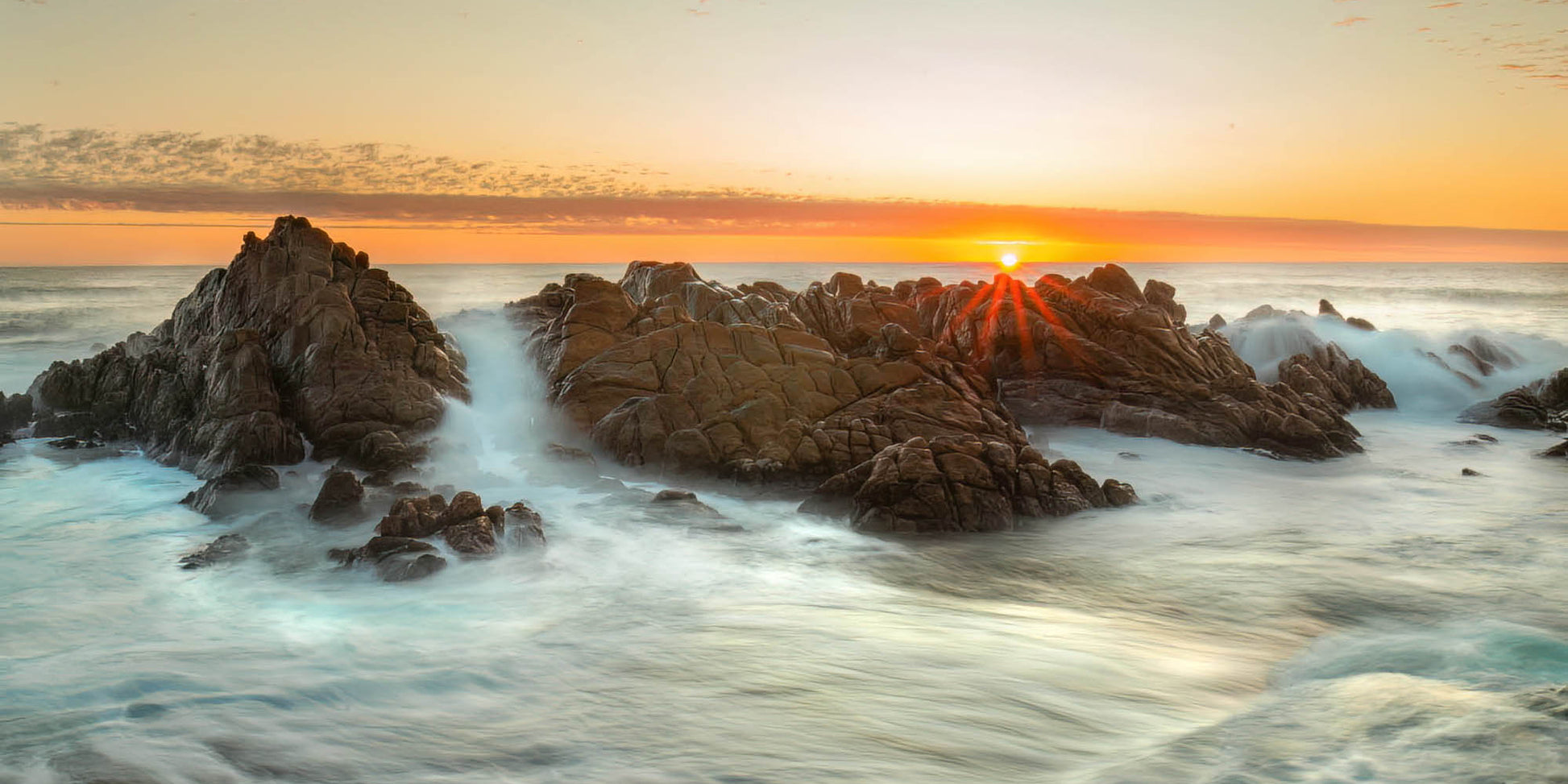 Sunset over rocky coastline with waves crashing against the rocks.