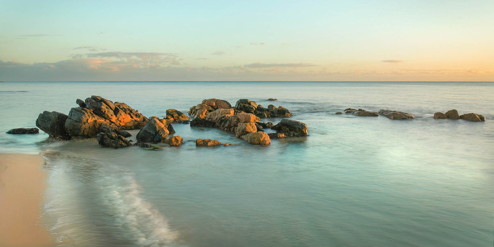 Rocks on a beach with a calm ocean and sky at sunrise.