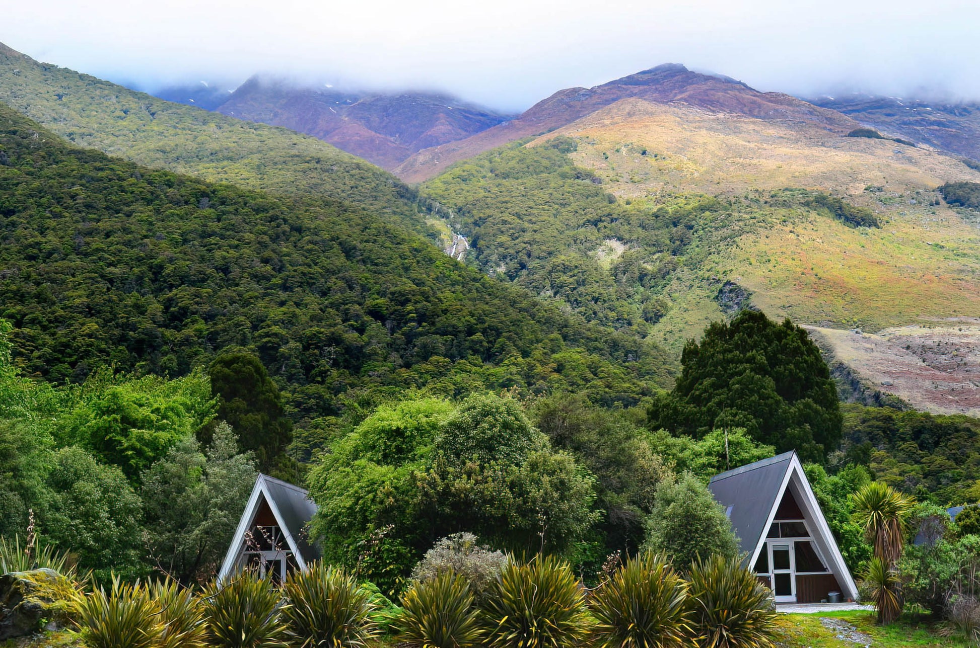 Two triangular cabins nestled among lush greenery with mountains in the background.