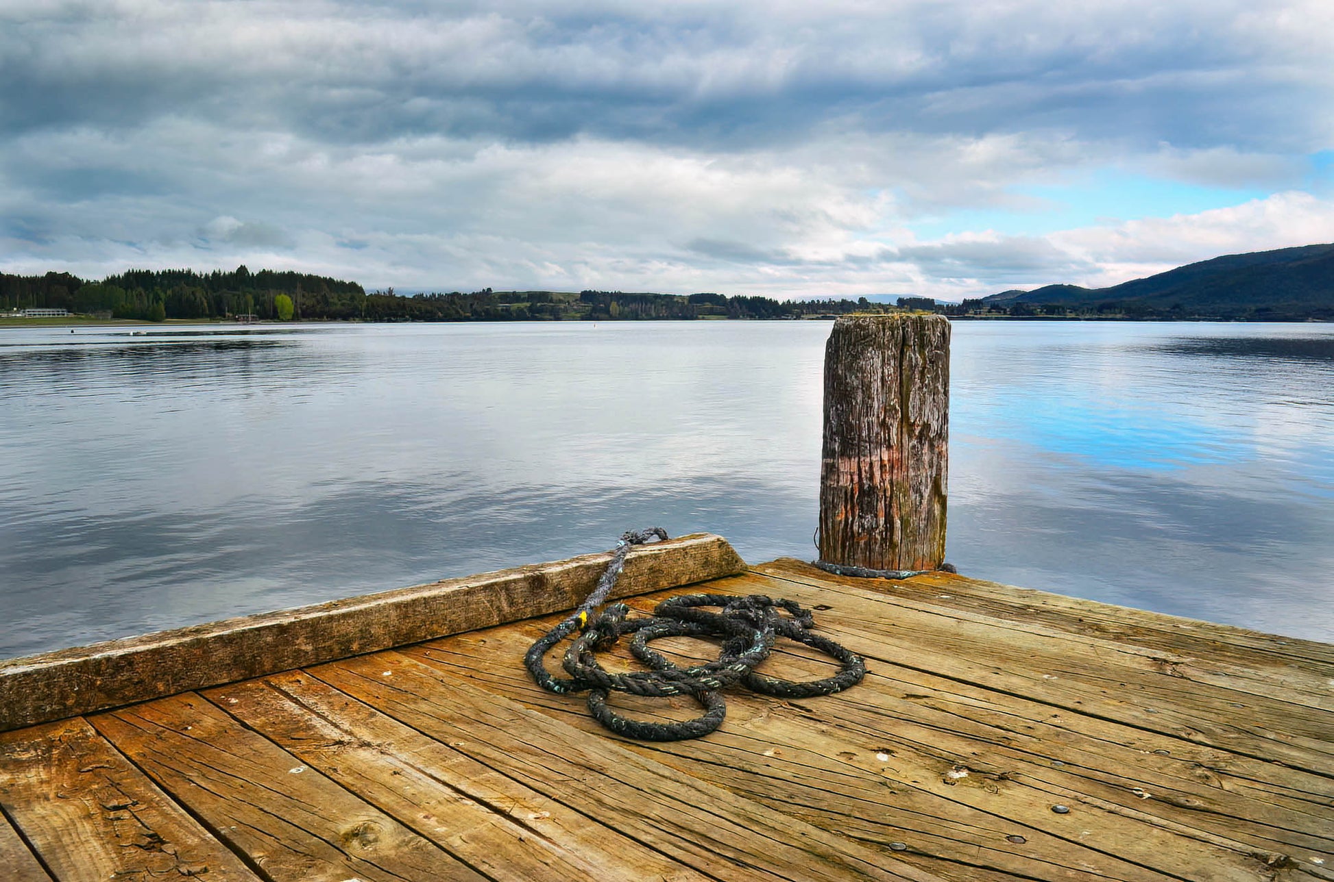 Wooden dock with a rope tied to a post, overlooking a calm lake with mountains in the distance.