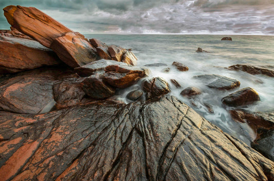 Rocks on a beach with waves crashing against them, under a cloudy sky.