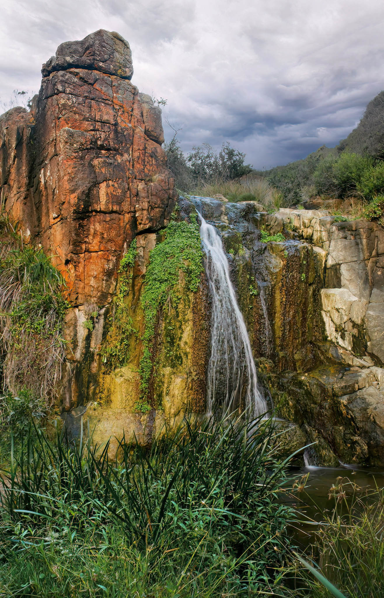 Waterfall cascading down rocky cliffs with greenery and a cloudy sky.