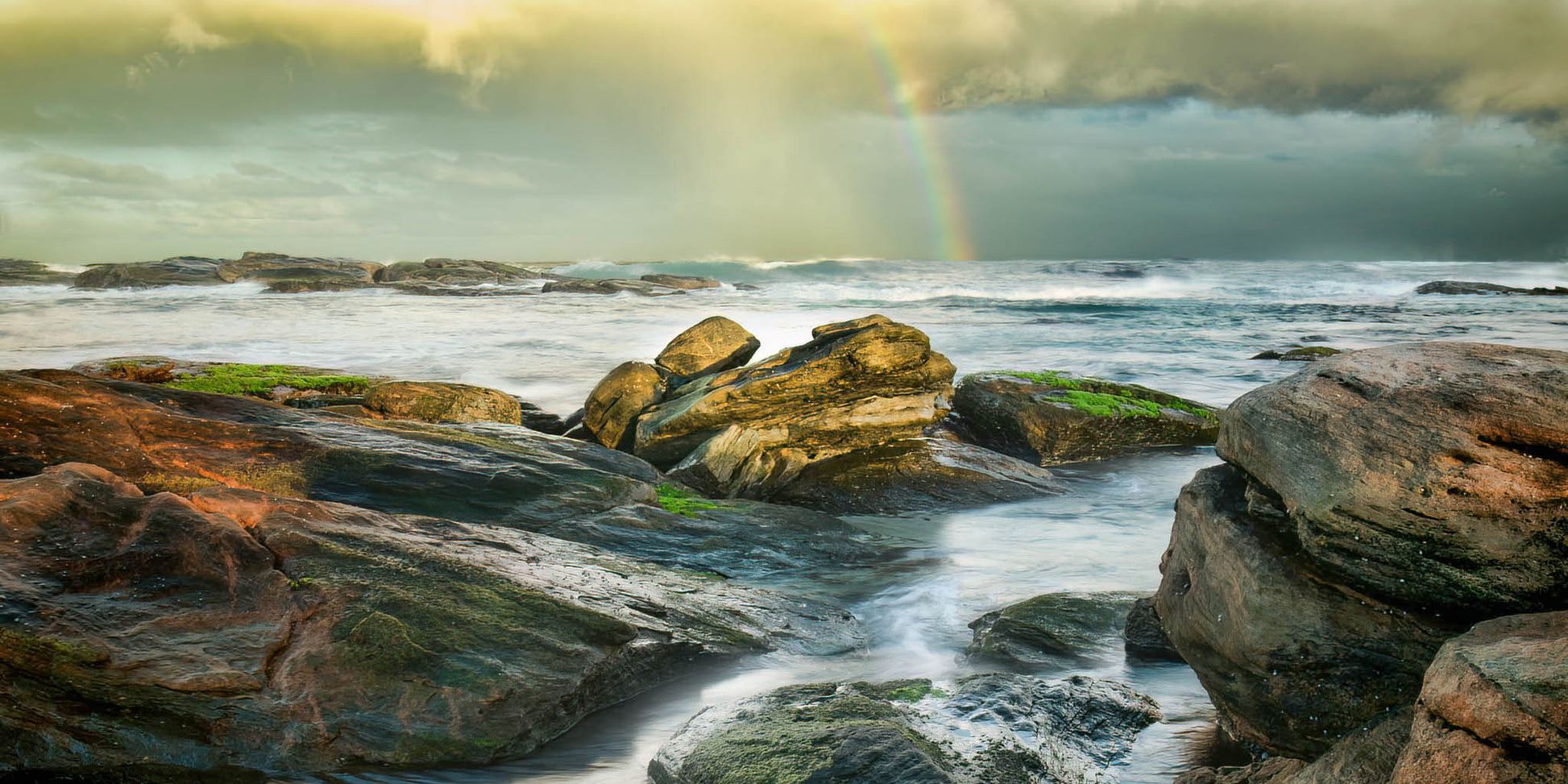 Rocks by the ocean with a rainbow in the background during a moody storm.