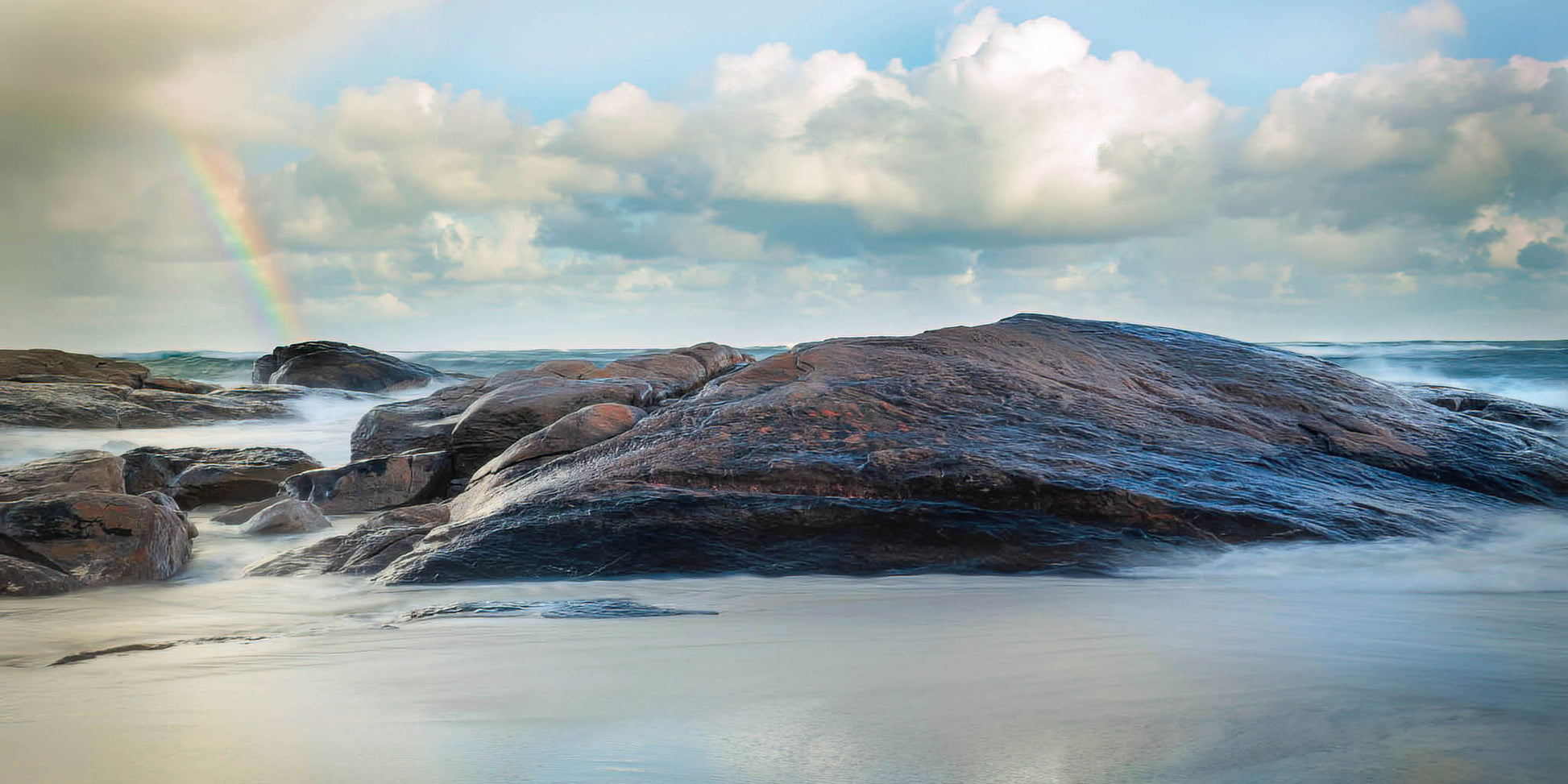 Rocks at Redgate Beach with a rainbow in a cloudy sky.