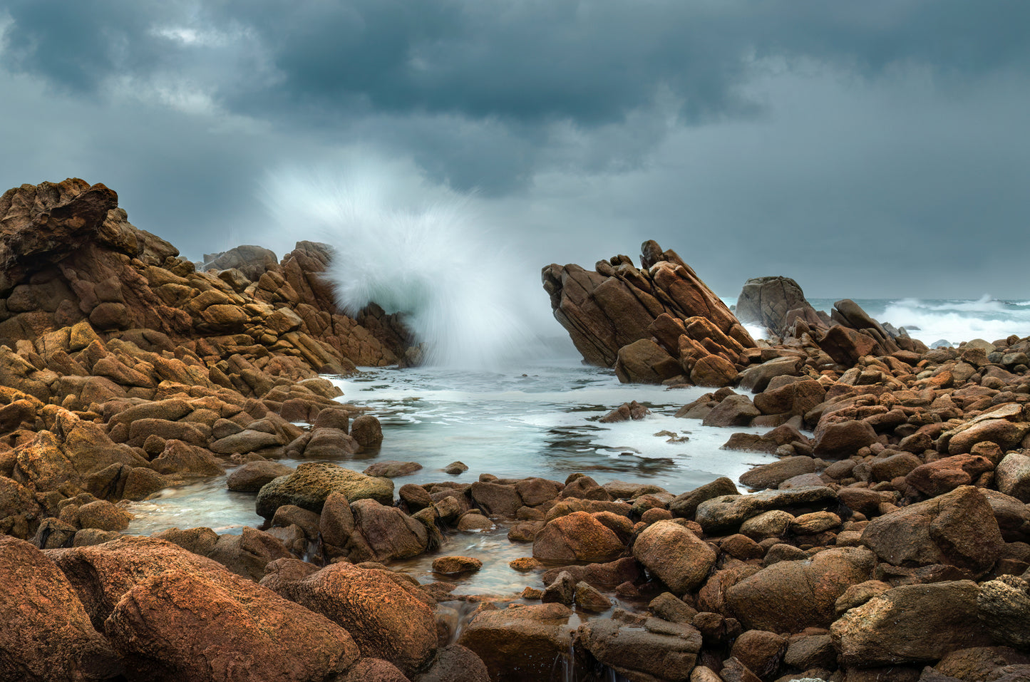 Long-exposure of wave explosion against rocks surrounding a calm rockpool.